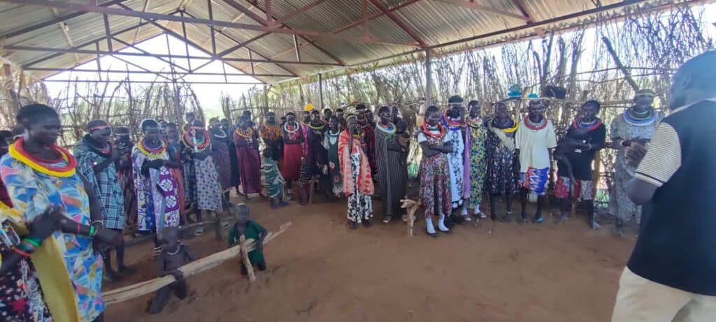 Community women gathered under a shelter in Kenya, wearing colorful traditional clothing and jewelry.
